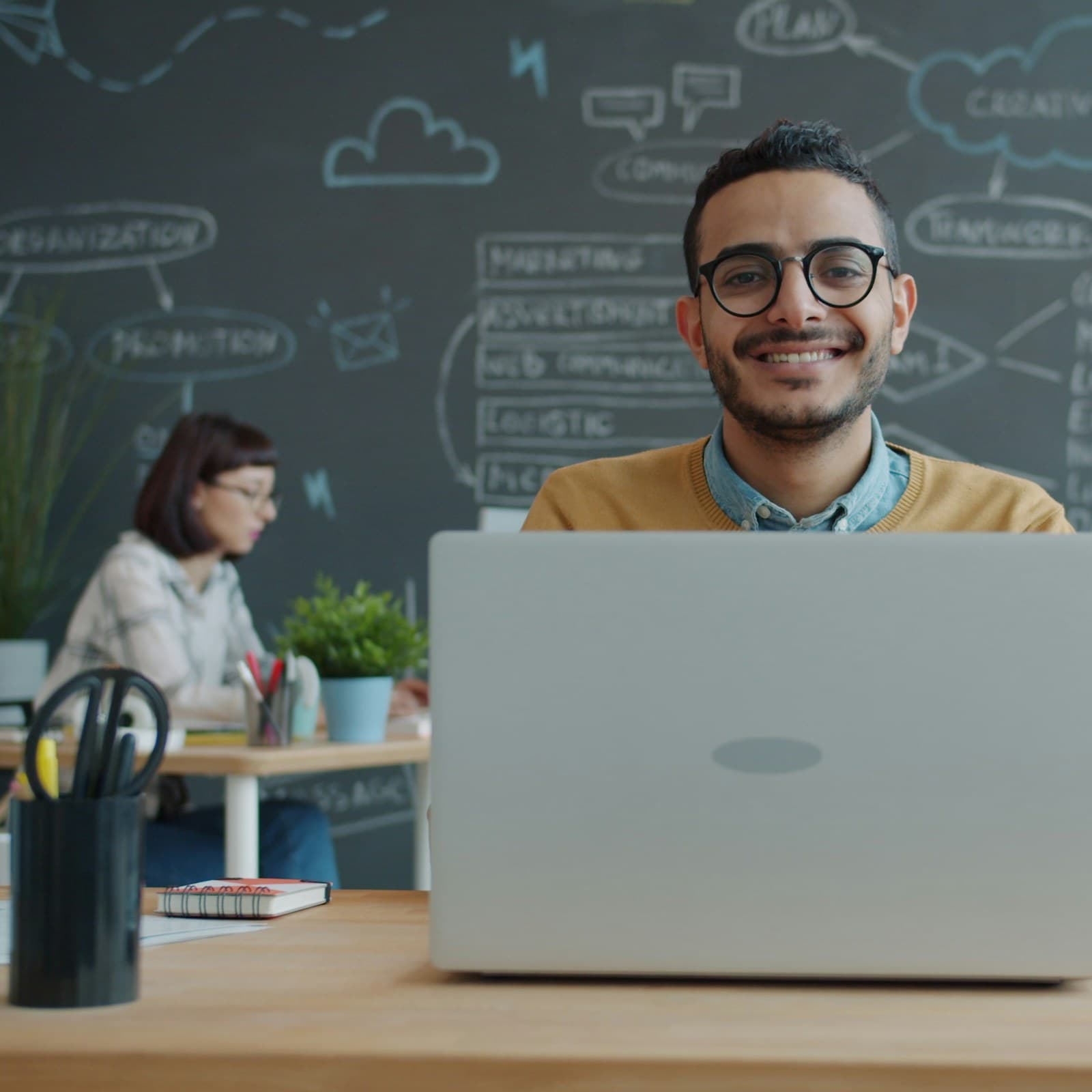A smiling man in glasses at his laptop, in an office with a strategy board behind him